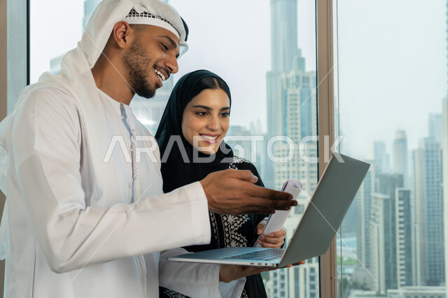 Developing and studying new projects, meetings and workshops at the company's headquarters, using modern technical devices, collecting data and solving problems, a close-up side view of an Arab Gulf Emirati businessman wearing a ghandoora and a ghutra discussing the project with his colleague