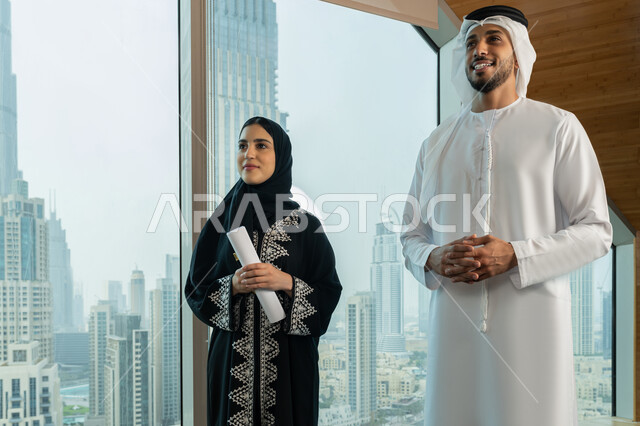 Data collection and problem solving, developing and studying new projects, comfortable work environment in UAE companies, meetings and workshops at company headquarters, a veiled Emirati Gulf Arab woman wearing an abaya holding a set of papers in her hand discussing the project with her colleague