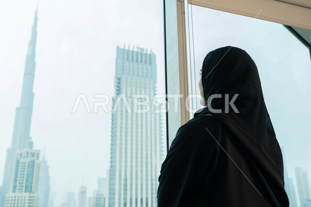 Concept of planning for the future, comfortable work environment in UAE companies, close-up from the back of a veiled Gulf Arab Emirati female employee wearing a black abaya looking out the window, glass facade overlooking towers and skyscrapers, elegant Saudi young woman