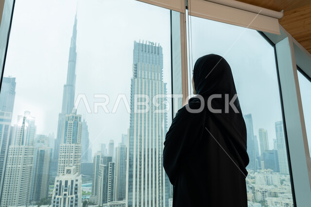 بيئة العمل المريحة في شركات الامارات، مفهوم التخطيط للمستقبل، صورة مقربة من الخلف لموظفة عربية خليجية اماراتية محجبة ترتدي عباءة سوداء تنظر عبر النافذة، واجهة زجاجية مطلة على الابراج وناطحات السحاب، شابة سعودية انيقة