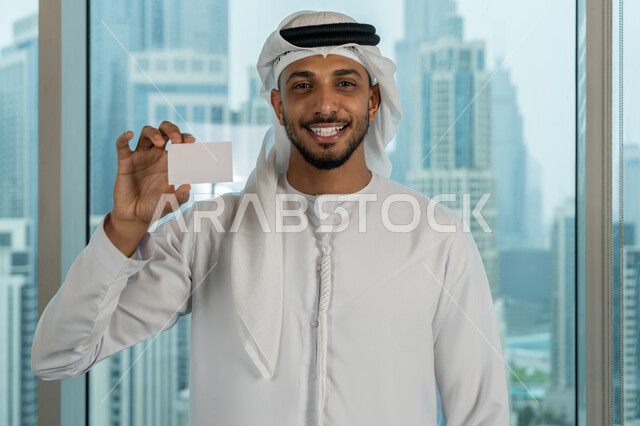 الوقوف باستقامة والنظر الى الكاميرا بايماءات السرور والسعادة، موك أب أبيض مستطيل فارغ، عرض بطاقة تعريفية، صورة مقربة لرجل عربي خليجي اماراتي يرتدي الكندورة والغترة يحمل بيده هوية شخصية، بيئة العمل المريحة في شركات الامارات