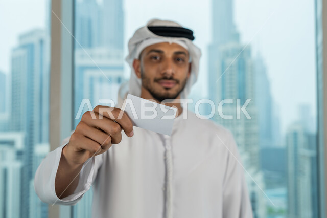 موك أب أبيض مستطيل فارغ، عرض بطاقة تعريفية، صورة مقربة لرجل عربي خليجي اماراتي يرتدي الكندورة والغترة يحمل بيده هوية شخصية، الوقوف باستقامة والنظر الى الكاميرا بايماءات السرور والسعادة، بيئة العمل المريحة في شركات الامارات