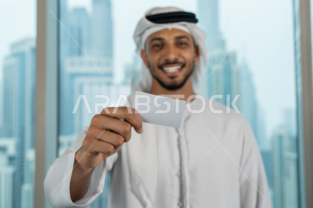 عرض بطاقة تعريفية، موك أب أبيض مستطيل فارغ، صورة مقربة لرجل عربي خليجي اماراتي يرتدي الكندورة والغترة يحمل بيده هوية شخصية، الوقوف باستقامة والنظر الى الكاميرا بايماءات السرور والسعادة، بيئة العمل المريحة في شركات الامارات