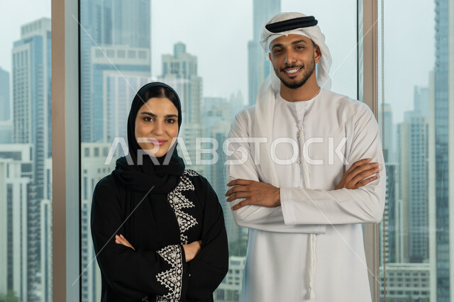 Standing straight and looking at the camera with gestures of self-confidence, understanding and cooperation between colleagues to achieve the company's goals, close-up of two Emirati Gulf Arab employees standing with crossed hands, comfortable work environment in UAE companies