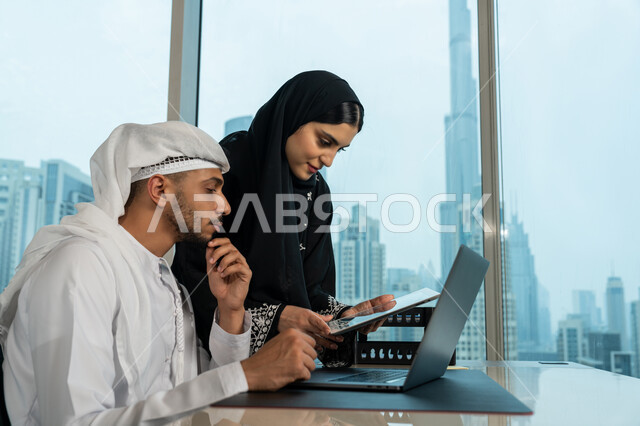 Data collection and problem solving, using modern technology devices, meetings and workshops at the company headquarters, a close-up side view of an Emirati Gulf Arab businessman wearing a ghandoora and a ghutra discussing the project with his colleague inside the office, developing and studying new projects