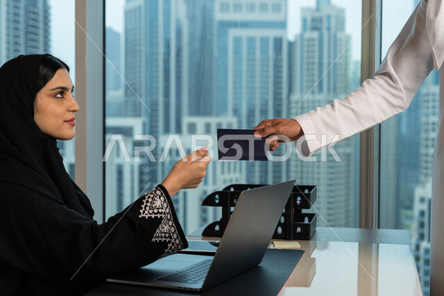 مفهوم حجز رحلات الطيران، نقل وتسجيل البيانات الشخصية، صورة مقربة ليد رجل عربي خليجي اماراتي يقوم بتمرير جواز السفر للموظفة، شابة سعودية محجبة تعمل على جهاز الحاسوب، بيئة العمل المريحة في شركات الامارات