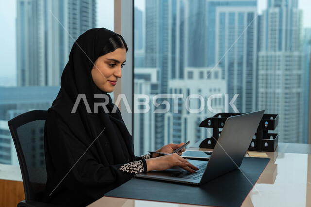 مفهوم حجز رحلات الطيران، نقل وتسجيل البيانات الشخصية، صورة مقربة لامرأة عربية خليجية اماراتية محجبة ترتدي العباءة السوداء تحمل بيدها جواز السفر، شابة سعودية تعمل على جهاز الحاسوب، بيئة العمل المريحة في شركات الامارات