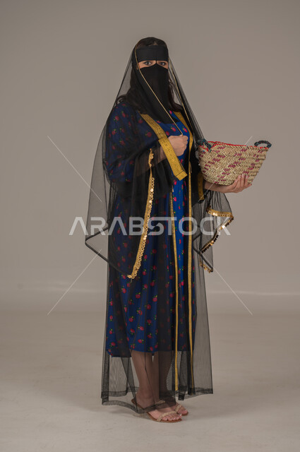 حرف ومهن يدوية شعبية, ارتداء زي المنطقة الوسطى في المناسبات الوطنية، يوم بدينا 1727م، ذكرى تأسيس الدولة السعودية الأولى 22 فبراير, الفخر بالتراث التاريخي، بورتريه لإمرأة عربية خليجية سعودية ترتدي البرقع والشيلة تحمل سلة قش, الجسم بالطول الكامل، خلفية رمادية