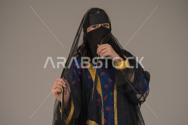 الفخر والاعتزاز بأنجازات البلاد، ارتداء زي المنطقة الوسطى في المناسبات الوطنية، يوم بدينا 1727 م، ذكرى تأسيس الدولة السعودية الأولى 22 فبراير, بورتريه مقرب لإمرأة عربية خليجية سعودية ترتدي البرقع والشيلة تنظر الى الكاميرا بتعابير الانوثة والنعومة، خلفية رمادية