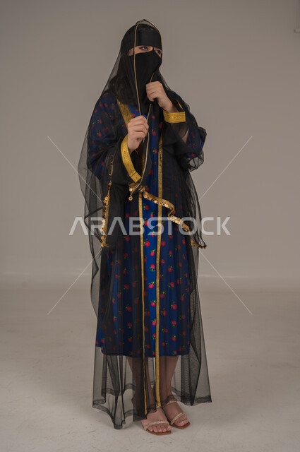 الفخر والاعتزاز بأنجازات البلاد، ارتداء زي المنطقة الوسطى في المناسبات الوطنية، يوم بدينا 1727 م، ذكرى تأسيس الدولة السعودية الأولى 22 فبراير, بورتريه لإمرأة عربية خليجية سعودية ترتدي البرقع والشيلة تقف بتعابير الانوثة والنعومة، صورة الجسم بالكامل, خلفية رمادية