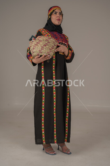 يوم بدينا 1727م, ارتداء زي المنطقة الوسطى في المناسبات الوطنية، ذكرى تأسيس الدولة السعودية الأولى 22 فبراير, مهن وحرف يدوية شعبية, الفخر بالتراث التاريخي، بورتريه لإمرأة عربية خليجية سعودية ترتدي الكرتة والهامة تحمل سلة من القش, الجسم بالطول الكامل، خلفية رمادية