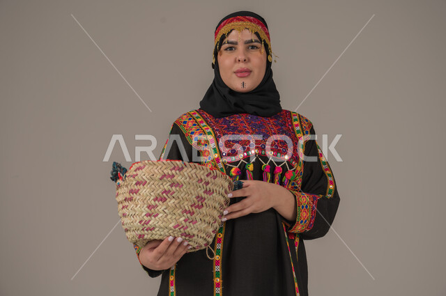 مهن وحرف يدوية شعبية, الفخر بالتراث التاريخي، بورتريه مقرب لإمرأة عربية خليجية سعودية ترتدي الكرتة والهامة تحمل سلة مصنوعة من القش, ارتداء زي المنطقة الوسطى في المناسبات الوطنية، يوم بدينا 1727م, ذكرى تأسيس الدولة السعودية الأولى في 22 فبراير, خلفية رمادية