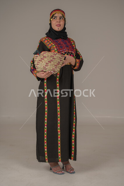 يوم بدينا 1727م, ارتداء زي المنطقة الوسطى في المناسبات الوطنية، ذكرى تأسيس الدولة السعودية الأولى 22 فبراير, مهن وحرف يدوية شعبية, الفخر بالتراث التاريخي، بورتريه لإمرأة عربية خليجية سعودية ترتدي الكرتة والهامة تحمل سلة من القش, الجسم بالطول الكامل، خلفية رمادية