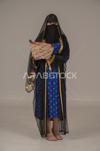 حرف ومهن يدوية شعبية, ارتداء زي المنطقة الوسطى في المناسبات الوطنية، يوم بدينا 1727م، ذكرى تأسيس الدولة السعودية الأولى 22 فبراير, الفخر بالتراث التاريخي، بورتريه لإمرأة عربية خليجية سعودية ترتدي البرقع والشيلة تحمل سلة قش, الجسم بالطول الكامل، خلفية رمادية