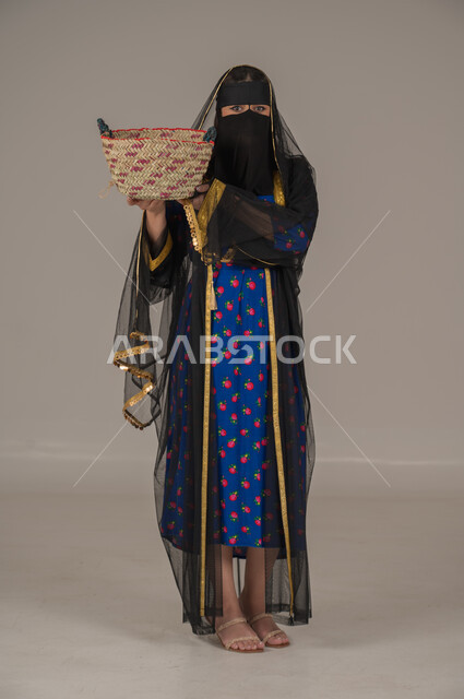 حرف ومهن يدوية شعبية, ارتداء زي المنطقة الوسطى في المناسبات الوطنية، يوم بدينا 1727م، ذكرى تأسيس الدولة السعودية الأولى 22 فبراير, الفخر بالتراث التاريخي، بورتريه لإمرأة عربية خليجية سعودية ترتدي البرقع والشيلة تحمل سلة قش, الجسم بالطول الكامل، خلفية رمادية
