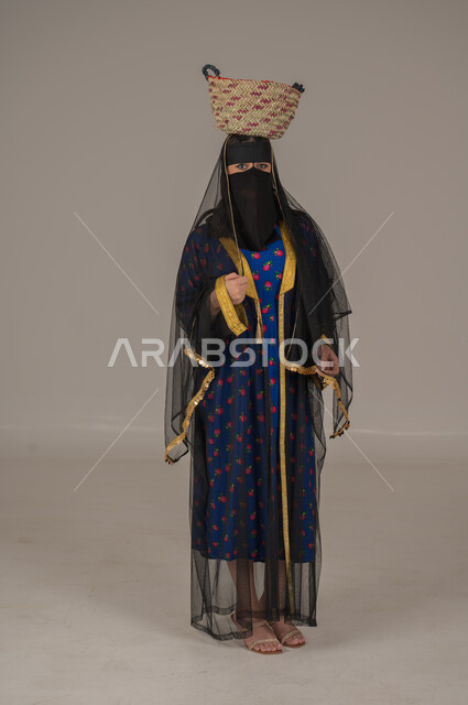 مهن يدوية شعبية, ارتداء زي المنطقة الوسطى في المناسبات الوطنية، يوم بدينا 1727م، ذكرى تأسيس الدولة السعودية الأولى 22 فبراير, الفخر بالتراث التاريخي، بورتريه لإمرأة عربية خليجية سعودية ترتدي البرقع والشيلة تضع  سلة قش على رأسها, الجسم بالطول الكامل، خلفية رمادية