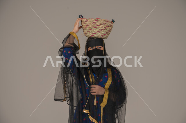 ذكرى تأسيس الدولة السعودية الأولى 22 فبراير, الفخر بالتراث التاريخي، مهن يدوية شعبية, ارتداء زي المنطقة الوسطى في المناسبات الوطنية، يوم بدينا 1727م، بورتريه مقرب لإمرأة عربية خليجية سعودية ترتدي البرقع والشيلة تضع سلة قش على رأسها، خلفية رمادية