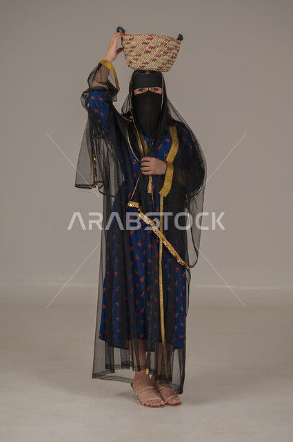 مهن يدوية شعبية, ارتداء زي المنطقة الوسطى في المناسبات الوطنية، يوم بدينا 1727م، ذكرى تأسيس الدولة السعودية الأولى 22 فبراير, الفخر بالتراث التاريخي، بورتريه لإمرأة عربية خليجية سعودية ترتدي البرقع والشيلة تضع  سلة قش على رأسها, الجسم بالطول الكامل، خلفية رمادية