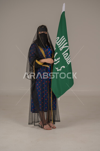 تأسيس الدولة السعودية الأولى 22 فبراير، ذكرى يوم بدينا 1727 م، ارتداء زي المنطقة الوسطى في المناسبات الشعبية والوطنية, بورتريه لإمرأة عربية خليجية سعودية ترتدي البرقع والشيلة تمسك راية وشعار يوم التأسيس بتعابير الفخر والاعتزاز, الجسم بالطول الكامل, خلفية رمادية