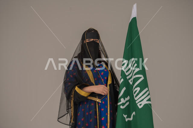 يوم بدينا 1727 م، ذكرى تأسيس الدولة السعودية الأولى 22 فبراير، ارتداء زي المنطقة الوسطى في المناسبات الشعبية والوطنية, بورتريه مقرب لإمرأة عربية خليجية سعودية ترتدي البرقع والشيلة تمسك راية وشعار يوم التأسيس بتعابير الفخر والاعتزاز, خلفية رمادية