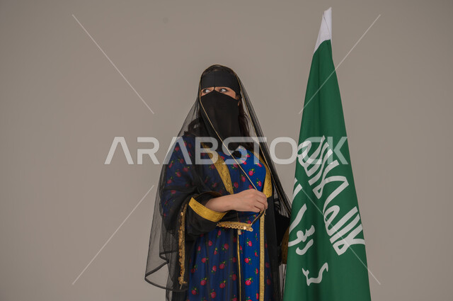 The Day We Began 1727 AD, the anniversary of the founding of the first Saudi state, February 22, wearing the Central Region costume on popular and national occasions, a close-up portrait of a Saudi Gulf Arab woman wearing a burqa and a shayla holding the banner and logo of the Founding Day with expressions of pride and honor, gray background