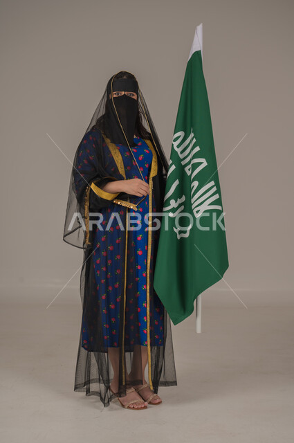 تأسيس الدولة السعودية الأولى 22 فبراير، ذكرى يوم بدينا 1727 م، ارتداء زي المنطقة الوسطى في المناسبات الشعبية والوطنية, بورتريه لإمرأة عربية خليجية سعودية ترتدي البرقع والشيلة تمسك راية وشعار يوم التأسيس بتعابير الفخر والاعتزاز, الجسم بالطول الكامل, خلفية رمادية