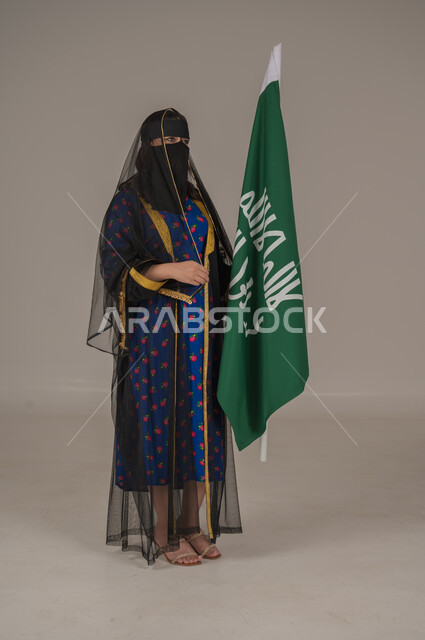 تأسيس الدولة السعودية الأولى 22 فبراير، ذكرى يوم بدينا 1727 م، ارتداء زي المنطقة الوسطى في المناسبات الشعبية والوطنية, بورتريه لإمرأة عربية خليجية سعودية ترتدي البرقع والشيلة تمسك راية وشعار يوم التأسيس بتعابير الفخر والاعتزاز, الجسم بالطول الكامل, خلفية رمادية