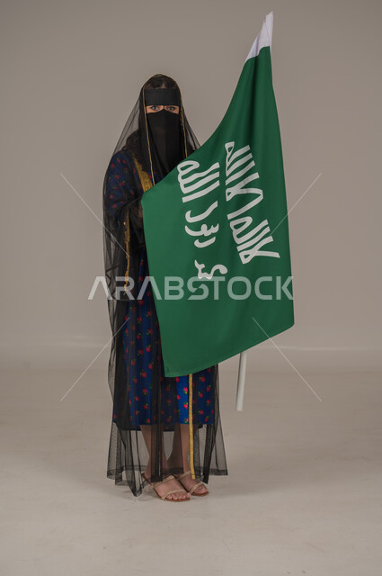 تأسيس الدولة السعودية الأولى 22 فبراير، ذكرى يوم بدينا 1727 م، ارتداء زي المنطقة الوسطى في المناسبات الشعبية والوطنية, بورتريه لإمرأة عربية خليجية سعودية ترتدي البرقع والشيلة تمسك راية وشعار يوم التأسيس بتعابير الفخر والاعتزاز, الجسم بالطول الكامل, خلفية رمادية