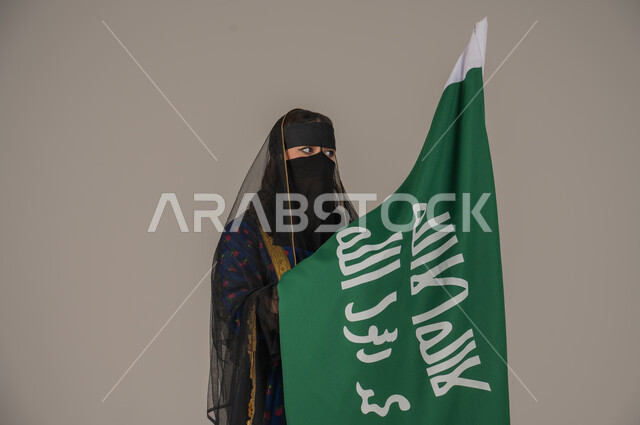 يوم بدينا 1727 م، ذكرى تأسيس الدولة السعودية الأولى 22 فبراير، ارتداء زي المنطقة الوسطى في المناسبات الشعبية والوطنية, بورتريه مقرب لإمرأة عربية خليجية سعودية ترتدي البرقع والشيلة تمسك راية وشعار يوم التأسيس بتعابير الفخر والاعتزاز, خلفية رمادية