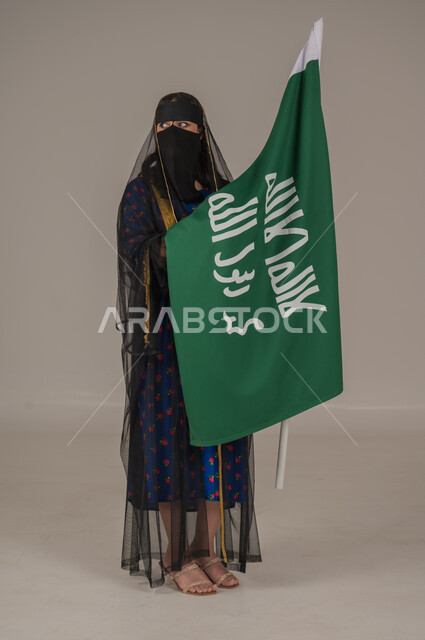 تأسيس الدولة السعودية الأولى 22 فبراير، ذكرى يوم بدينا 1727 م، ارتداء زي المنطقة الوسطى في المناسبات الشعبية والوطنية, بورتريه لإمرأة عربية خليجية سعودية ترتدي البرقع والشيلة تمسك راية وشعار يوم التأسيس بتعابير الفخر والاعتزاز, الجسم بالطول الكامل, خلفية رمادية