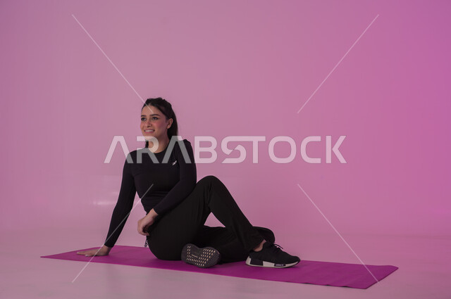 نمط حياة صحي ومفيد، ممارسة التمرينات الأرضية لشد الجسد، مفهوم الرشاقة واللياقة، انشطة  بدنية نسائية، تقوية العضلات ونحت الجسم، المحافظة على التوازن الجسدي، بورتريه لشابة عربية خليجية سعودية ترتدي زي رياضي أسود تفوم بممارسة التمرينات الرياضية ،إيماءات الحماس و النشاط، خلفية وردية