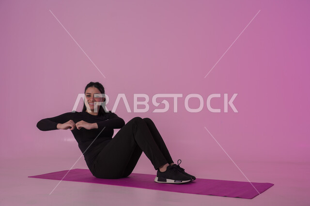 نمط حياة صحي ومفيد، ممارسة التمرينات الأرضية لشد الجسد، مفهوم الرشاقة واللياقة، انشطة  بدنية نسائية، تقوية العضلات ونحت الجسم، المحافظة على التوازن الجسدي، بورتريه لشابة عربية خليجية سعودية ترتدي زي رياضي أسود تفوم بممارسة التمرينات الرياضية ،إيماءات الحماس و النشاط، خلفية وردية