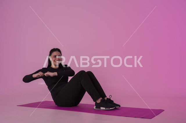 نمط حياة صحي ومفيد، ممارسة التمرينات الأرضية لشد الجسد، مفهوم الرشاقة واللياقة، انشطة  بدنية نسائية، تقوية العضلات ونحت الجسم، المحافظة على التوازن الجسدي، بورتريه لشابة عربية خليجية سعودية ترتدي زي رياضي أسود تفوم بممارسة التمرينات الرياضية ،إيماءات الحماس و النشاط، خلفية وردية