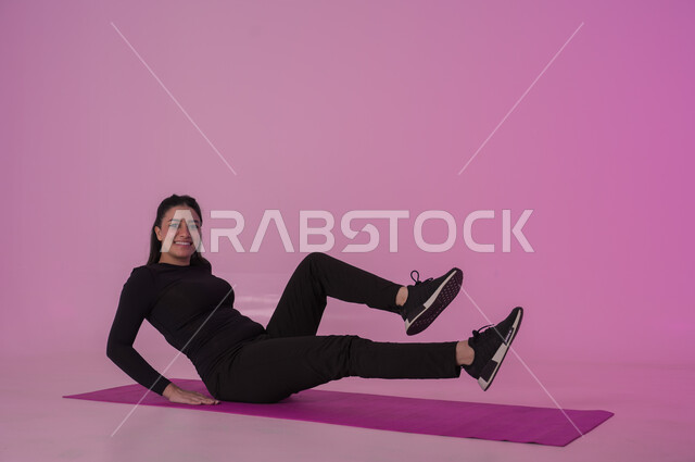 نمط حياة صحي ومفيد، ممارسة التمرينات الأرضية لشد الجسد، مفهوم الرشاقة واللياقة، انشطة  بدنية نسائية، تقوية العضلات ونحت الجسم، المحافظة على التوازن الجسدي، بورتريه لشابة عربية خليجية سعودية ترتدي زي رياضي أسود تفوم بممارسة التمرينات الرياضية ،إيماءات الحماس و النشاط، خلفية وردية