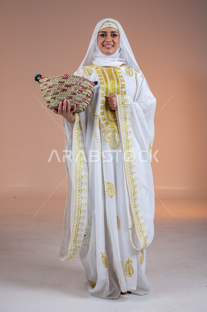 مهن وحرف يدوية شعبية قديمة، بورتريه لامرأة عربية خليجية سعودية مبتسمة ترتدي الشيلة والنشل تمسك بيدها سلة مصنوعة من القش، النظر إلى الكاميرا بتعابير الفخر والاعتزاز، يوم بدينا١٧٢٧م، ارتداء أزياء المنطقة الشرقية، ذكرى تأسيس الدولة السعودية الأولى٢٢فبراير، خلفية بنية