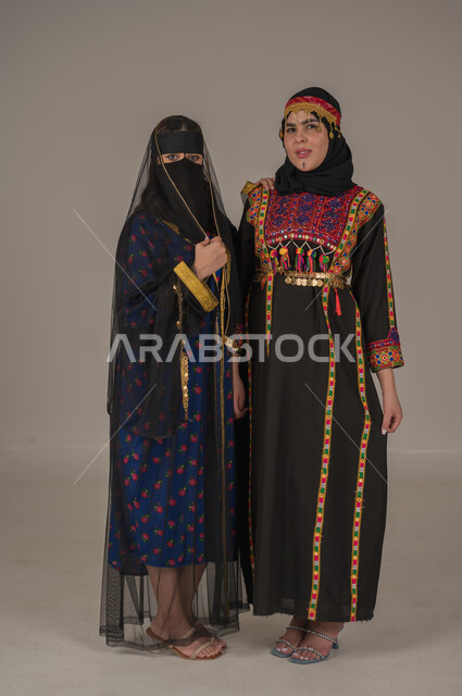 النظر إلى الكاميرا بإيماءات الفخر والاعتزاز، ارتداء الأزياء التقليدية للمنطقة الوسطى، الاحتفال بالمناسبات والأعياد الوطنية، بورتريه لامرأتين عربيتين خليجيتين سعوديتين ترتديان زي يوم تأسيس الدولة السعودية الاولى 22 فبراير، يوم بدينا ١٧٢٧م, صورة الجسم الكامل بالطول، خلفية رمادية