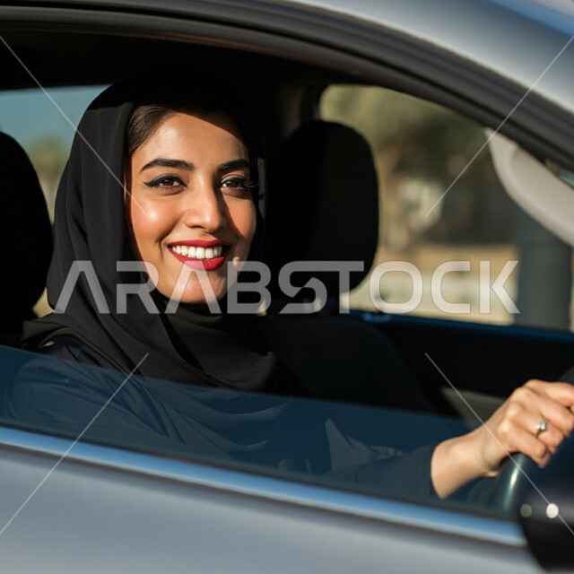 القيادة بحذر واتباع قواعد السلامة، قضاء وقت ممتع في الخارج، إيماءات السرور والسعادة، صورة مقربة من الجانب لامرأة عربيه خليجية سعودية مبتسمة ترتدي الحجاب والعباءة تجلس في مقعد السيارة، مفهوم الاستقلال والحرية والاعتماد على النفس