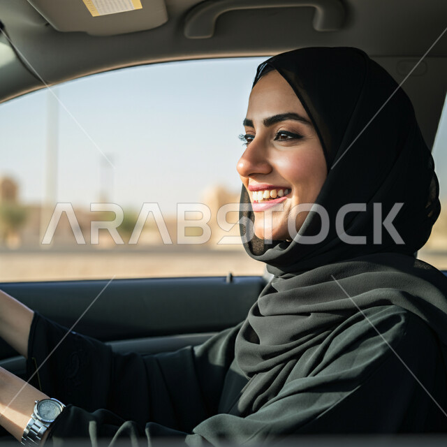 القيادة بحذر واتباع قواعد السلامة، قضاء وقت ممتع في الخارج، إيماءات السرور والسعادة، صورة مقربة من الجانب لامرأة عربيه خليجية سعودية مبتسمة ترتدي الحجاب والعباءة تجلس في مقعد السيارة، مفهوم الاستقلال والحرية والاعتماد على النفس