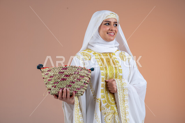 النظر إلى مكان ما بتعابير الفخر والاعتزاز، يوم بدينا١٧٢٧م، مهن وحرف يدوية شعبية قديمة، بورتريه لامرأة عربية خليجية سعودية مبتسمة ترتدي الشيلة والنشل تمسك بيدها سلة قش، ارتداء أزياء المنطقة الشرقية، ذكرى تأسيس الدولة السعودية الأولى٢٢فبراير، خلفية بنية