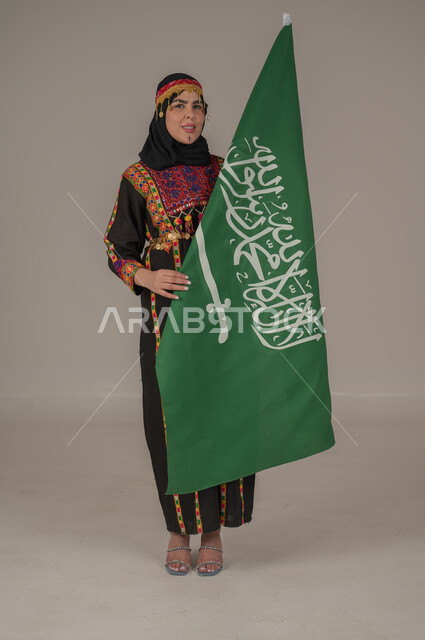 الاحتفال بالمناسبات الوطنية, يوم بدينا ١٧٢٧م، بورتريه لإمرأة عربيه سعودية خليجية ترتدي الهامة والشيلة والكرتة تحمل بيدها علم المملكة، ذكرى تأسيس الدولة السعودية الأولى، يوم العلم السعودي 11 مارس, إرتداء أزياء المنطقة الوسطى, الجسم الكامل بالطول، خلفيه رمادية