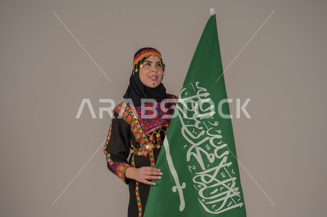 تعابير الفخر والاعتزاز، الاحتفال بالمناسبات الوطنية, يوم بدينا ١٧٢٧م، بورتريه لإمرأة عربيه سعودية خليجية ترتدي الهامة والشيلة والكرتة تحمل بيدها علم المملكة، ذكرى تأسيس الدولة السعودية الأولى، يوم العلم السعودي 11 مارس, إرتداء أزياء المنطقة الوسطى، خلفيه رمادية