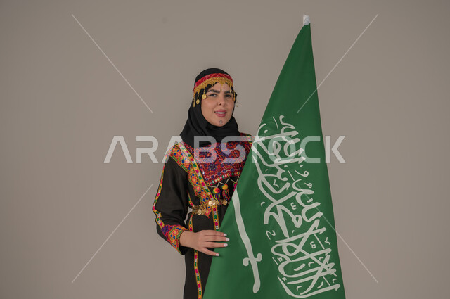تعابير الفخر والاعتزاز، الاحتفال بالمناسبات الوطنية, يوم بدينا ١٧٢٧م، بورتريه لإمرأة عربيه سعودية خليجية ترتدي الهامة والشيلة والكرتة تحمل بيدها علم المملكة، ذكرى تأسيس الدولة السعودية الأولى، يوم العلم السعودي 11 مارس, إرتداء أزياء المنطقة الوسطى، خلفيه رمادية