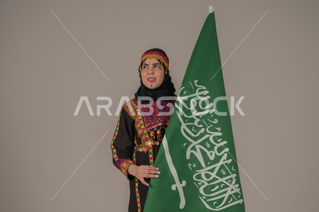 تعابير الفخر والاعتزاز، الاحتفال بالمناسبات الوطنية, يوم بدينا ١٧٢٧م، بورتريه لإمرأة عربيه سعودية خليجية ترتدي الهامة والشيلة والكرتة تحمل بيدها علم المملكة، ذكرى تأسيس الدولة السعودية الأولى، يوم العلم السعودي 11 مارس, إرتداء أزياء المنطقة الوسطى، خلفيه رمادية