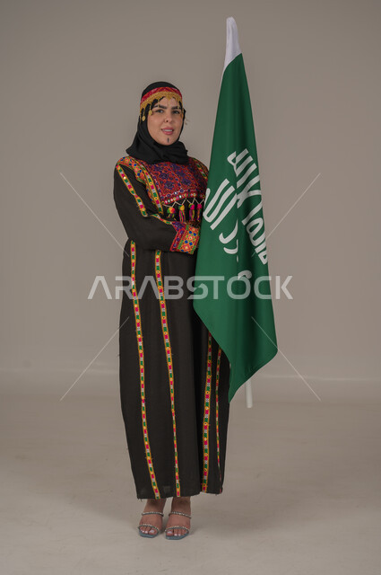 ذكرى تأسيس الدولة السعودية الأولى الاحتفال بالاعياد الوطنية, يوم بدينا ١٧٢٧م، بورتريه لإمرأة عربيه سعودية خليجية ترتدي الهامة والشيلة والكرتة تحمل بيدها علم يوم التأسيس، يوم العلم السعودي 11 مارس, إرتداء أزياء المنطقة الوسطى, الجسم الكامل بالطول، خلفيه رمادية