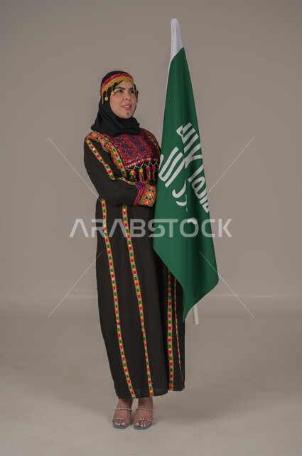ذكرى تأسيس الدولة السعودية الأولى الاحتفال بالاعياد الوطنية, يوم بدينا ١٧٢٧م، بورتريه لإمرأة عربيه سعودية خليجية ترتدي الهامة والشيلة والكرتة تحمل بيدها علم يوم التأسيس، يوم العلم السعودي 11 مارس, إرتداء أزياء المنطقة الوسطى, الجسم الكامل بالطول، خلفيه رمادية