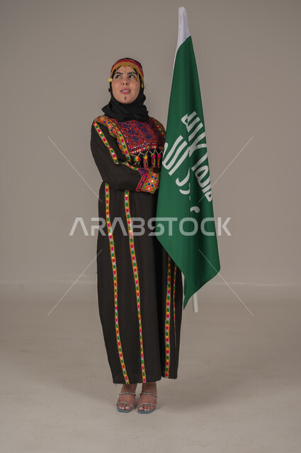 ذكرى تأسيس الدولة السعودية الأولى الاحتفال بالاعياد الوطنية, يوم بدينا ١٧٢٧م، بورتريه لإمرأة عربيه سعودية خليجية ترتدي الهامة والشيلة والكرتة تحمل بيدها علم يوم التأسيس، يوم العلم السعودي 11 مارس, إرتداء أزياء المنطقة الوسطى, الجسم الكامل بالطول، خلفيه رمادية