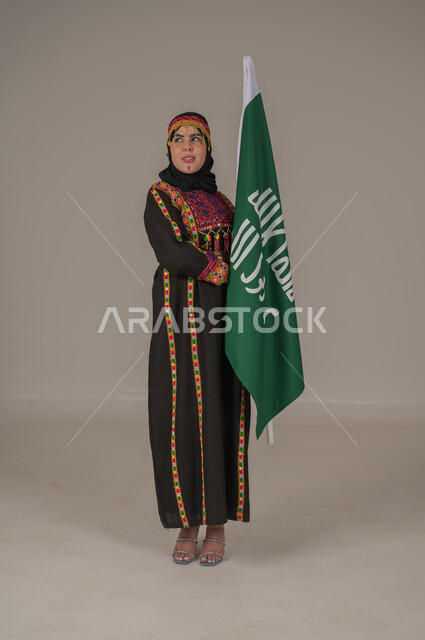 ذكرى تأسيس الدولة السعودية الأولى الاحتفال بالاعياد الوطنية, يوم بدينا ١٧٢٧م، بورتريه لإمرأة عربيه سعودية خليجية ترتدي الهامة والشيلة والكرتة تحمل بيدها علم يوم التأسيس، يوم العلم السعودي 11 مارس, إرتداء أزياء المنطقة الوسطى, الجسم الكامل بالطول، خلفيه رمادية