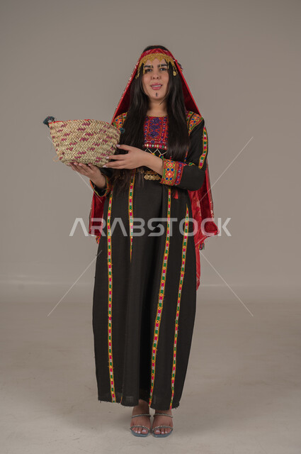  النظر الى الكاميرا بتعابير الفخر والاعتزاز، ارتداء أزياء المنطقة الوسطى, مهن وحرف يدوية شعبية، يوم بدينا ١٧٢٧م، بورتريه لإمرأه عربيه سعوديه خليجيه ترتدي الهامة والكرتة تمسك بيدها سلة قش، ذكرى تأسيس الدولة السعودية الأولى٢٢ فبراير، الجسم الكامل بالطول، خلفيه رمادية