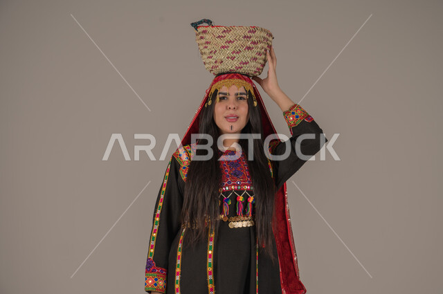 مهن وحرف يدوية شعبية تقليدية، يوم بدينا ١٧٢٧م، بورتريه لإمرأه عربيه سعوديه خليجيه ترتدي الهامة والكرتة تضع على رأسها سلة قش، النظر الى الكاميرا بتعابير الفخر والاعتزاز، ذكرى تأسيس الدولة السعودية الأولى٢٢ فبراير, ارتداء الازياء التقليدية للمنطقة الوسطى, خلفيه رمادية