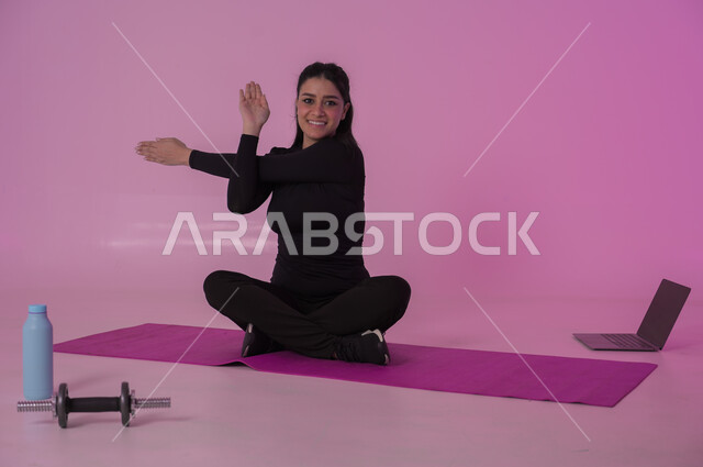 إيماءات السعادة والنشاط، أنشطة  بدنية للمحافظة على اللياقة والرشاقة، اتباع أسلوب حياة صحي باستخدام الأجهزة التقنية، رياضات لتقوية العضلات ونحت الجسم، بورتريه لشابة عربية خليجية سعودية ترتدي زي رياضي تقوم بمتابعة التمارين الرياضية عبر الحاسب المحمول، خلفية وردية